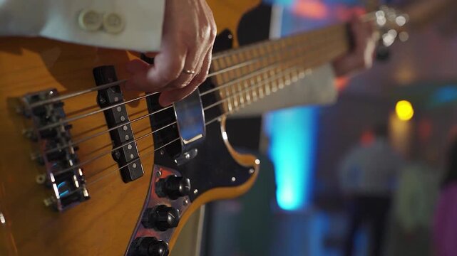 Cover band member, bassist, plays yellow bass guitar during party. Close-up of guitarist's hands in white jacket. People dancing in the background. Nightlife, live music at music festival.
