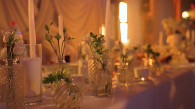 Bouquets of white roses stand in crystal bowls on table in wedding banquet hall against backdrop of flickering spotlights and hall lighting. Concept of style in field of organizing holidays, events.