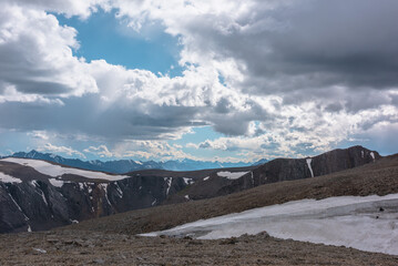 High stony pass, glacier, big rocky ridge, sheer crags and large mountain range with snowy pointy peaks far away under blue pure opening among clouds. Snow peaked tops under clear gap in cloudy sky. © Daniil