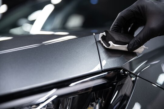 Close-up of a professional detailer applying ceramic or graphene coating to a car surface using an applicator. The process enhances gloss, provides hydrophobic properties