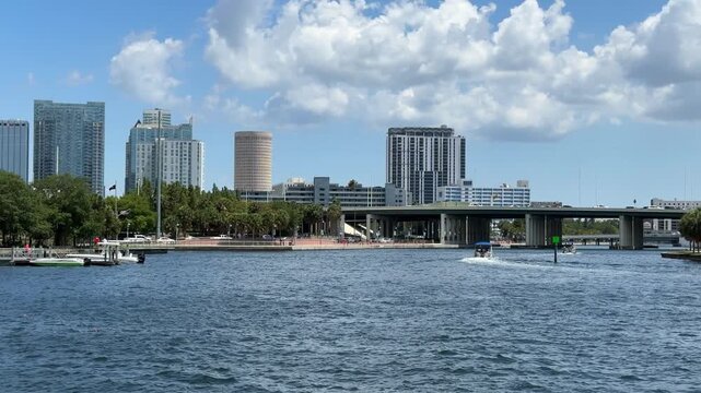 Skyscrapers and urban buildings stand behind a lush green shoreline along a sparkling waterway with boats. A large bridge spans the river under a bright blue sky filled with white puffy clouds.
