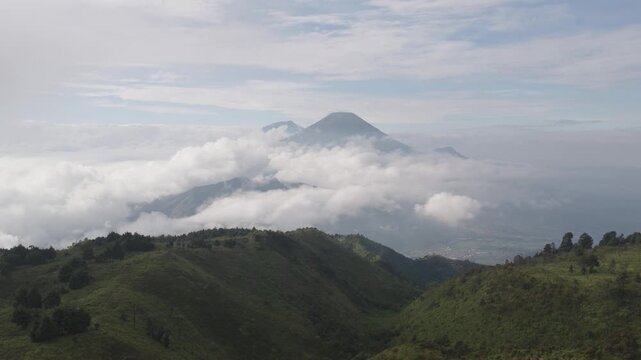 Aerial view of Gunung Prau peak rising above thick white clouds and lush green mountain ridges in Bawang, Central Java, Indonesia.