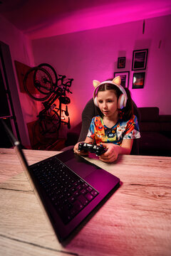 Young girl talking while playing online video games using laptop and controller in neon room