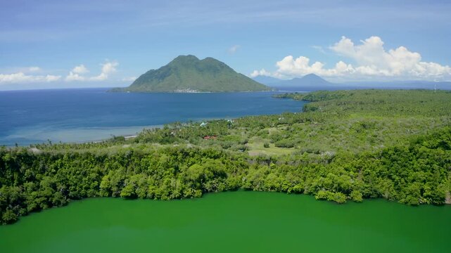 Aerial view of the vibrant green waters of a volcanic lake surrounded by lush tropical forest with a volcano across the sea in Ternate, North Maluku, Indonesia.