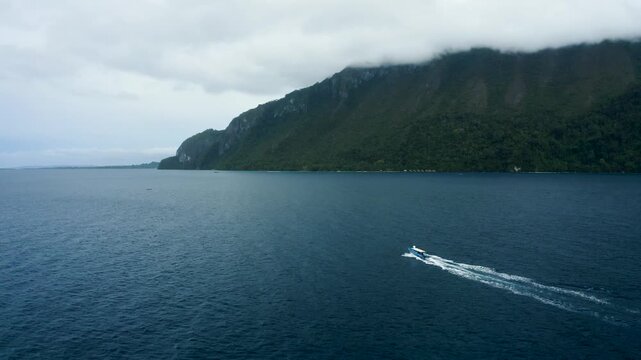 Aerial view of a white boat cruising through deep blue waters along the lush green cliffs of Seram Island Seram, Indonesia.