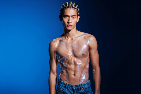Young man with braided hair wears a translucent PVC garment that catches sharp specular highlights against a cobalt blue backdrop. High-contrast studio lighting defines his musculature through the