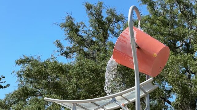 A giant orange bucket releases a cascade of water onto an outdoor aquatic playground. The water feature is surrounded by green trees under a clear blue sky.