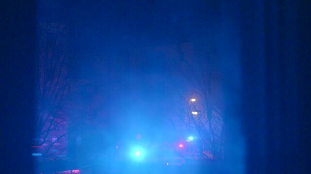 Blue and red lights flash over wet street, emergency vehicles converge near building facade, headlights cut through mist, responders secure scene with caution signs and traffic