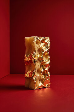 Crinkled gold foil tightly adheres to the porous surface of a rectangular sponge, creating sharp specular highlights against a matte crimson backdrop and deep red shadows.
