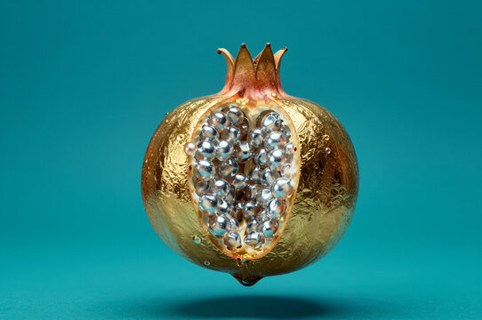 Split pomegranate features a textured metallic gold rind containing highly reflective liquid mercury droplets instead of seeds. Harsh top lighting creates specular highlights on the silver globules