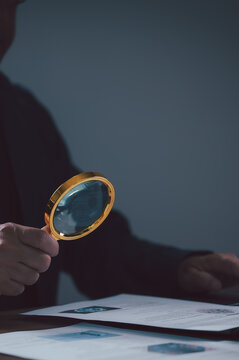 Professional investigator holds golden magnifying glass over paper document examine fine print details during private audit session inside dark office environment under dim light in evening time.