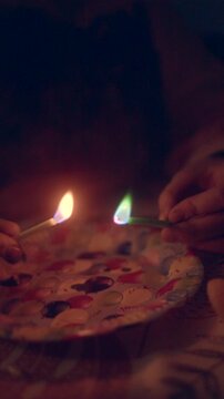 Hands lighting dual color matches over painted ceramic plate, orange and green flames meet above decorative dish, close composition shows fingers and flame detail, warm tactile