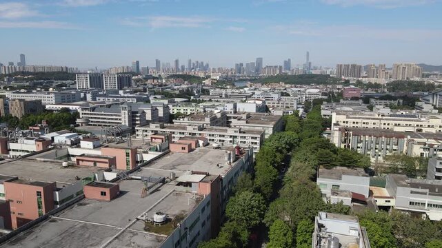 Aerial view of industrial factory buildings and lush green trees with the modern city skyline under a clear blue sky in Dongguan, Guangdong Province, China.
