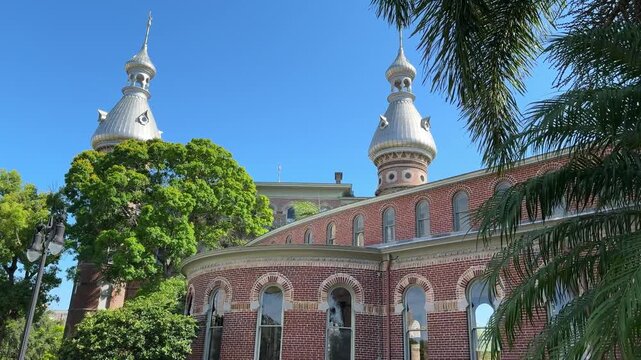 A circular wing of the historic university building displays classic Victorian brickwork and large arched windows. Lush green shrubs and palm trees surround the paved courtyard on a sunny Florida day.