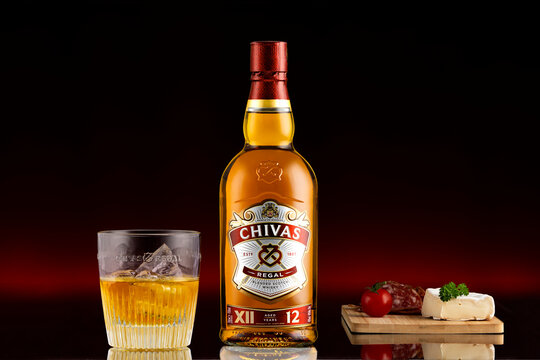Chivas Regal 12 years blended Scotch whisky bottle with glass and ice, presented with cheese and cold cuts on dark red background, illustrative editorial luxury alcohol concept.