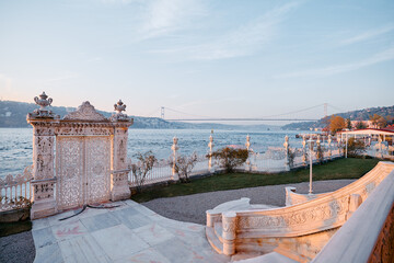 Fototapeta premium Elegan gates frame a waterfront view during sunset, with a bridge spanning the distant horizon and serene surroundings of greenery and architecture creating a picturesque and tranquil landscape.