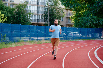 Fototapeta premium A man jogs on a red athletic track with an urban backdrop on a sunny day. Fitness and active lifestyle in a city setting, emphasizing health and exercise in everyday routines.