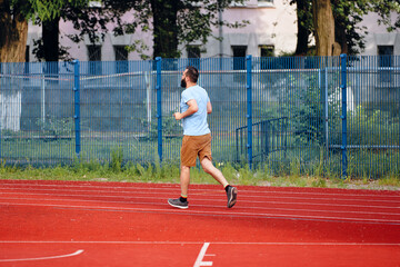 Obraz premium A man is running on an athletic track in a park area, wearing casual clothing. The image illustrates outdoor exercise, physical activity, and an active lifestyle in an urban environment.