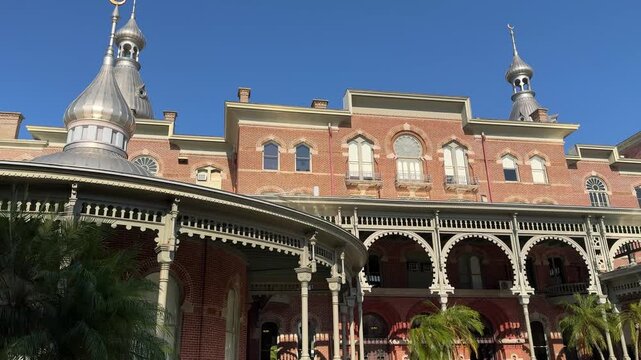 A decorative white wooden veranda curves around the exterior of a Victorian era landmark surrounded by lush palm trees. The red brick facade and silver minarets of the historic structure stand out