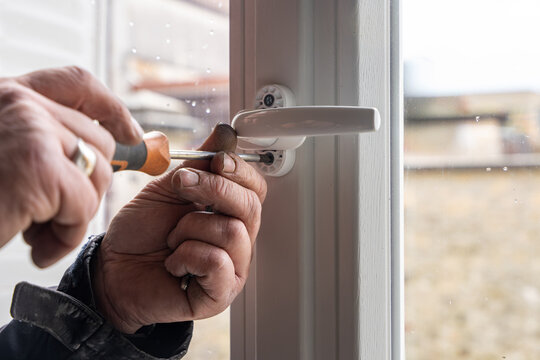 Gros plan sur les mains d'un artisan utilisant un tournevis pour ajuster avec pr&eacute;cision la poign&eacute;e d'une fen&ecirc;tre en PVC blanc lors d'une installation de menuiserie.