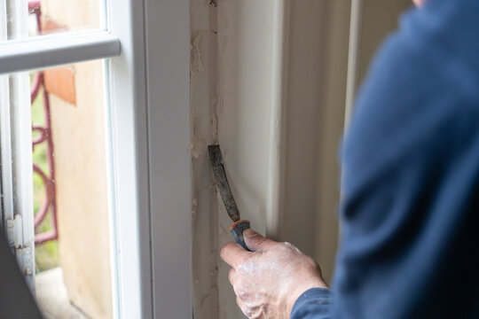Gros plan sur un travail de pr&eacute;paration minutieux : un artisan en bleu de travail gratte soigneusement l'ancien rev&ecirc;tement d'un &eacute;coin&ccedil;on de fen&ecirc;tre blanche avec une spatule m&eacute;tallique avant r&eacute;novation
