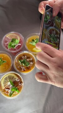Vertical stock video smartphone photographing drinks overhead. Hands of content creator styling iced beverages in plastic cups with lemon slices and mint leaves, ice cubes