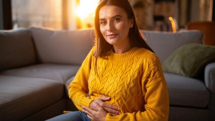 Obraz premium A woman with long brown hair sits on a couch wearing a yellow sweater, clutching her stomach in apparent discomfort or pain, set against a warm and cozy living room background.