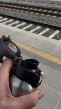 Hand holding water bottle near tracks, closeup of stainless sipper with flip lid, tactile line and rails in background, commuter pausing to hydrate while waiting, vertical stock