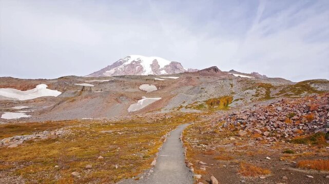A scenic view of the trail leading to Panorama Point, with the snow-capped peak of Mount Rainier dominating the background. The alpine meadows display vibrant orange and yellow fall colors under a par