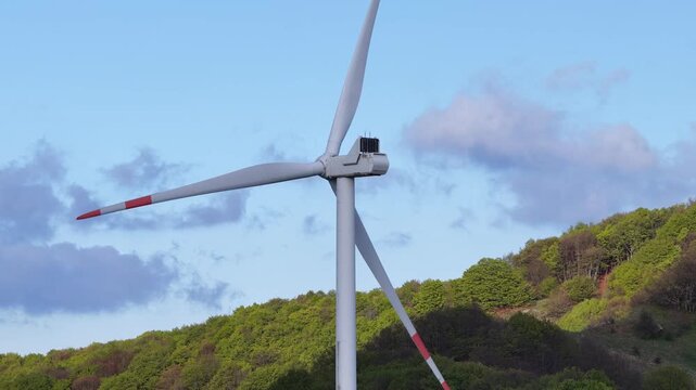 Aerial view of a wind turbine at Passo Di Cento Croci surrounded by green forested hills under a blue sky in Varese Ligure, Liguria, Italy.