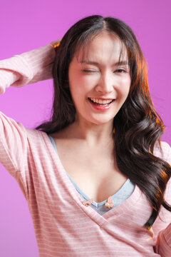 beautiful young East Asian woman with long wavy dark hair and a bright toothy smile, looking away and laughing. She is wearing a soft pink striped top with a light blue inner layer, set against a vibr