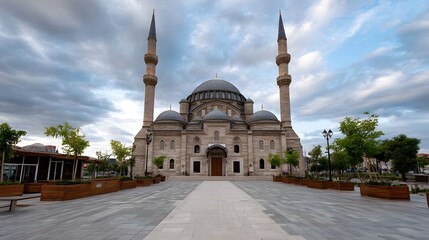 Fototapeta premium A majestic religious structure featuring domes and minarets dominates a wide plaza framed by lush greenery beneath a dramatic clouded sky