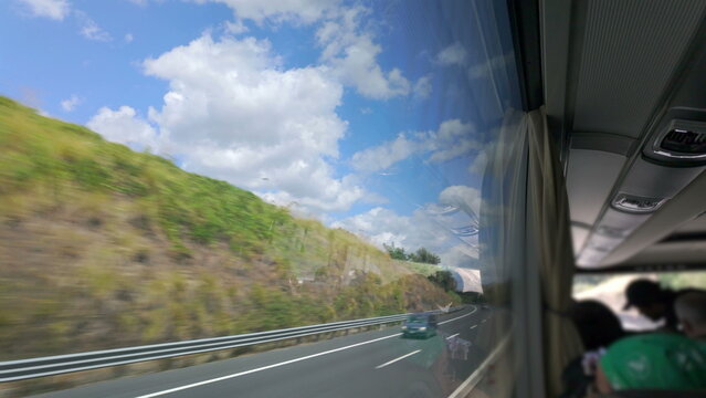 View from inside bus shows bright landscape with winding road, guardrail, and fluffy clouds outside, reflected in bus window next to passengers