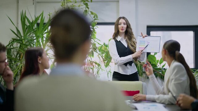 A confident businesswoman presents financial data and charts to her diverse team during an office meeting.