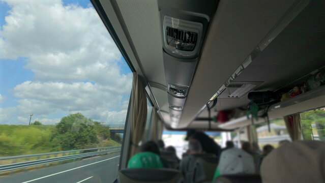 Interior view of moving tour bus with visible ceiling vents, curtains, and seated passengers, blue sky and highway bridge seen through wide window