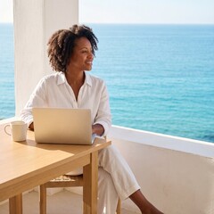Obraz premium A happy African American woman works remotely on her laptop from a sunny balcony overlooking the beautiful blue ocean, enjoying the freedom of a digital nomad lifestyle in a luxury resort