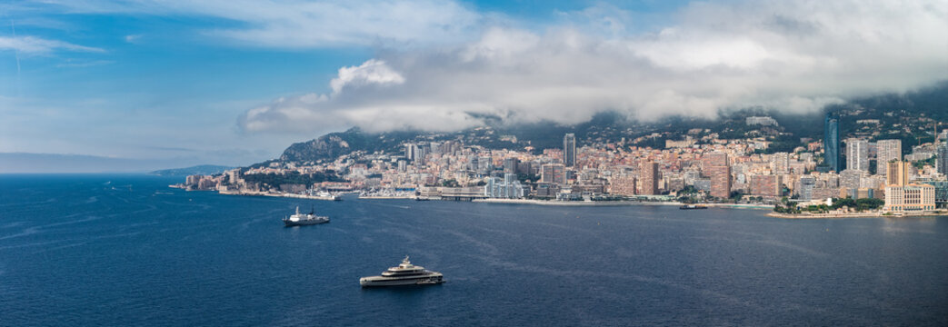 A wide aerial panorama shows Monaco on the French Riviera, with high rises and terraced blocks around a deep bay as yachts cruise and anchor under low clouded hills.