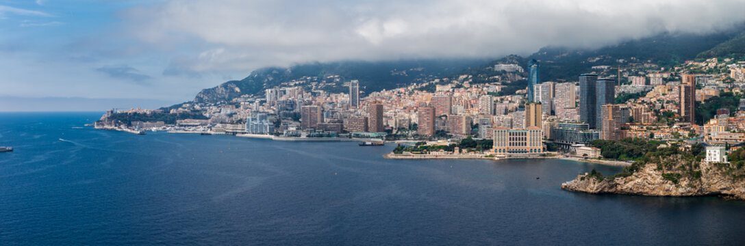 Aerial view of Monaco with Port Hercule and Monte Carlo, curved harbor walls, rocky headlands, high rise towers, terraced apartments, and cloud covered hills along the coast.