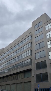 Vertical stock video urban office building under overcast sky concrete facade with grid windows reflecting clouds, van and parking sign at street level, towering commercial block,