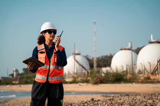 A confident female environmental engineer wearing full safety gear looks directly at the camera while working in a petrochemical refinery industrial setting.
