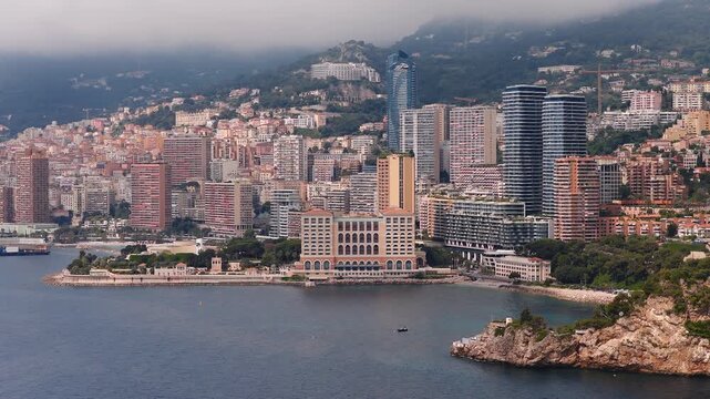 Aerial daylight pan shows Monaco high rises, arched beige waterfront hotel, rocky headland, blue gray sea, lone service boat, and misty hills near Monte Carlo.