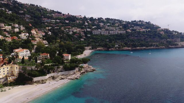 Aerial glide along Monaco Riviera shows villa dotted hills, Belle Epoque villa, pebble beach, yachts, swimmers, sunbathers, seawalls, limestone, soft daytime light.