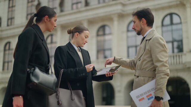 Business professionals conduct an outdoor meeting, discussing important documents and data on a tablet. Three corporate colleagues collaborate and communicate in an urban environment.