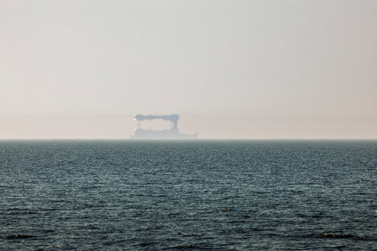 Sea view with ship Fata Morgana mirage on distant horizon in haze, atmospheric coastal scene with optical illusion and shimmering light over calm water
