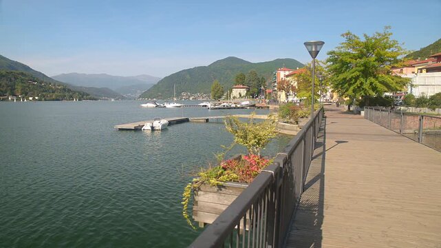 Lake Lugano summer panorama at Porto Ceresio with waterfront promenade, piers and boats, alpine mountains, clear blue sky, calm water, Italy Switzerland border, no people, tranquil scenic landscape