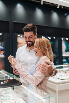 A happy couple examines jewelry in a high-end boutique. The man embraces the woman as they look at a display case.