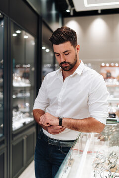A handsome young man in a white shirt tries on an expensive timepiece while standing in a luxury jewelry shop.