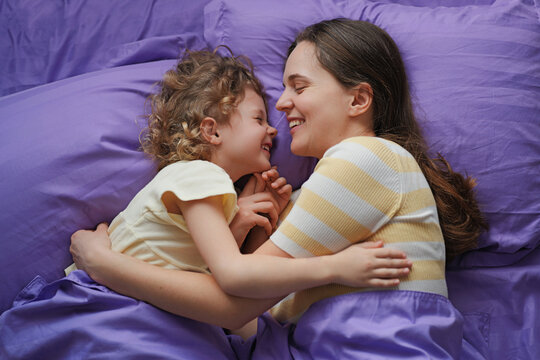 A mother and her child are lying close on a bed with purple sheets, sharing smiles and laughter. They enjoy a relaxed moment together at home in the afternoon