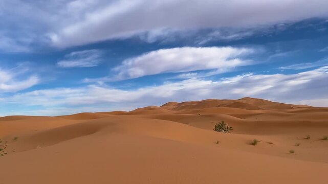Golden sand dunes stretch across the landscape, illuminated by warm sunlight under a clear blue sky and scattered clouds.