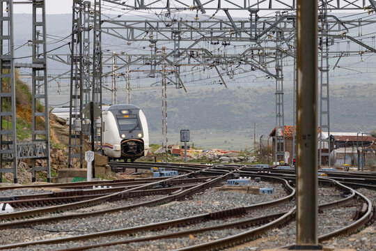 Commuters and travellers gather along the platforms at &Aacute;vila railway station as a regional train arrives, framed by overhead cables and urban architecture